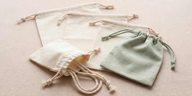 Five plain cotton drawstring bags on pale linen — a small open pouch with braided cords at the front, flat dust bags fanned behind and one sage-dyed pouch with a matching cord