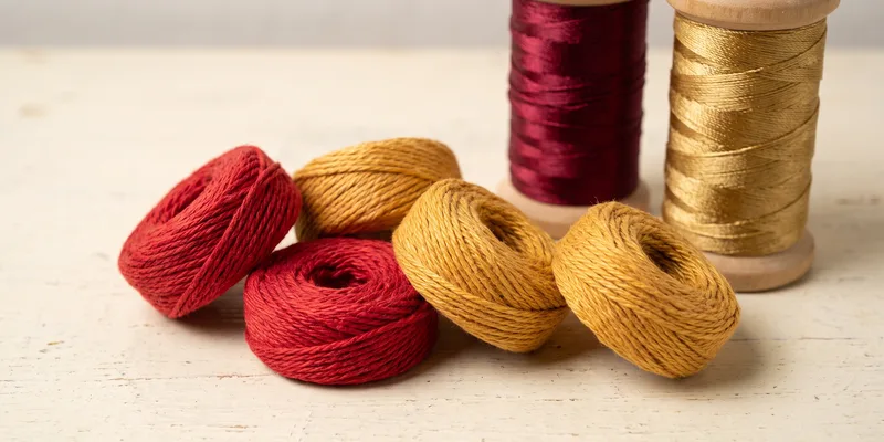 Flat rolls of red and of yellow twisted thread on a pale surface, with two wooden spools of glossy deep-red and gold rakhi-making thread standing behind