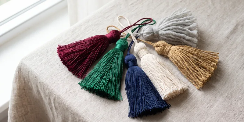 Six tassels fanned on pale linen — burgundy, emerald and navy rayon, natural cotton, golden jute and grey wool, each on its own cord loop