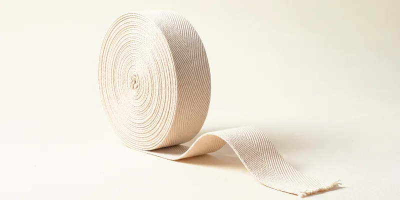 A roll of natural undyed cotton webbing standing on edge with its herringbone weave visible, and a length unwound in front ending in a frayed cut edge